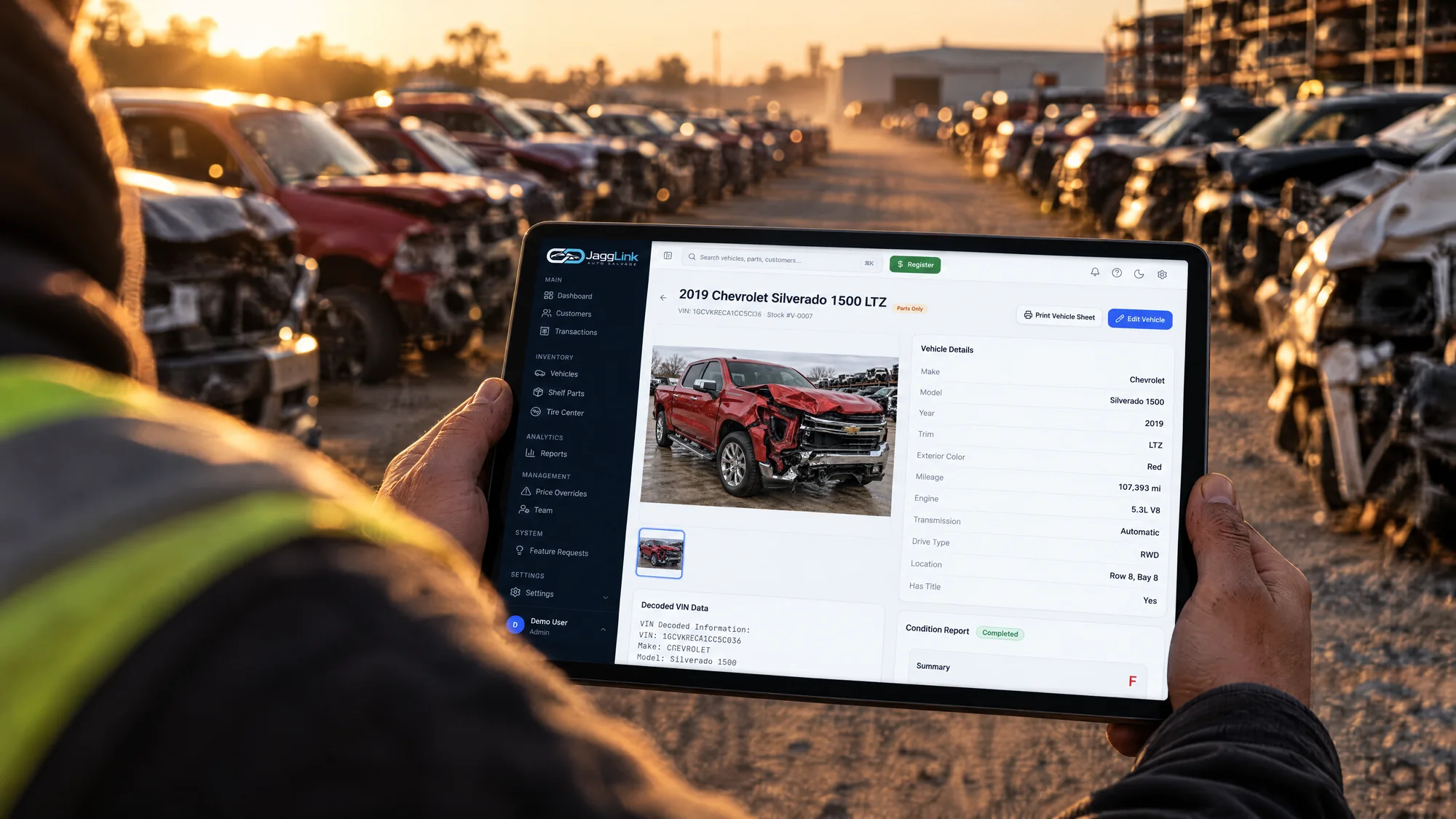 A salvage yard worker walking a row of wrecked vehicles, holding a tablet showing the JaggLink vehicle record for a 2019 Chevrolet Silverado 1500 LTZ with its decoded VIN, condition report and yard location